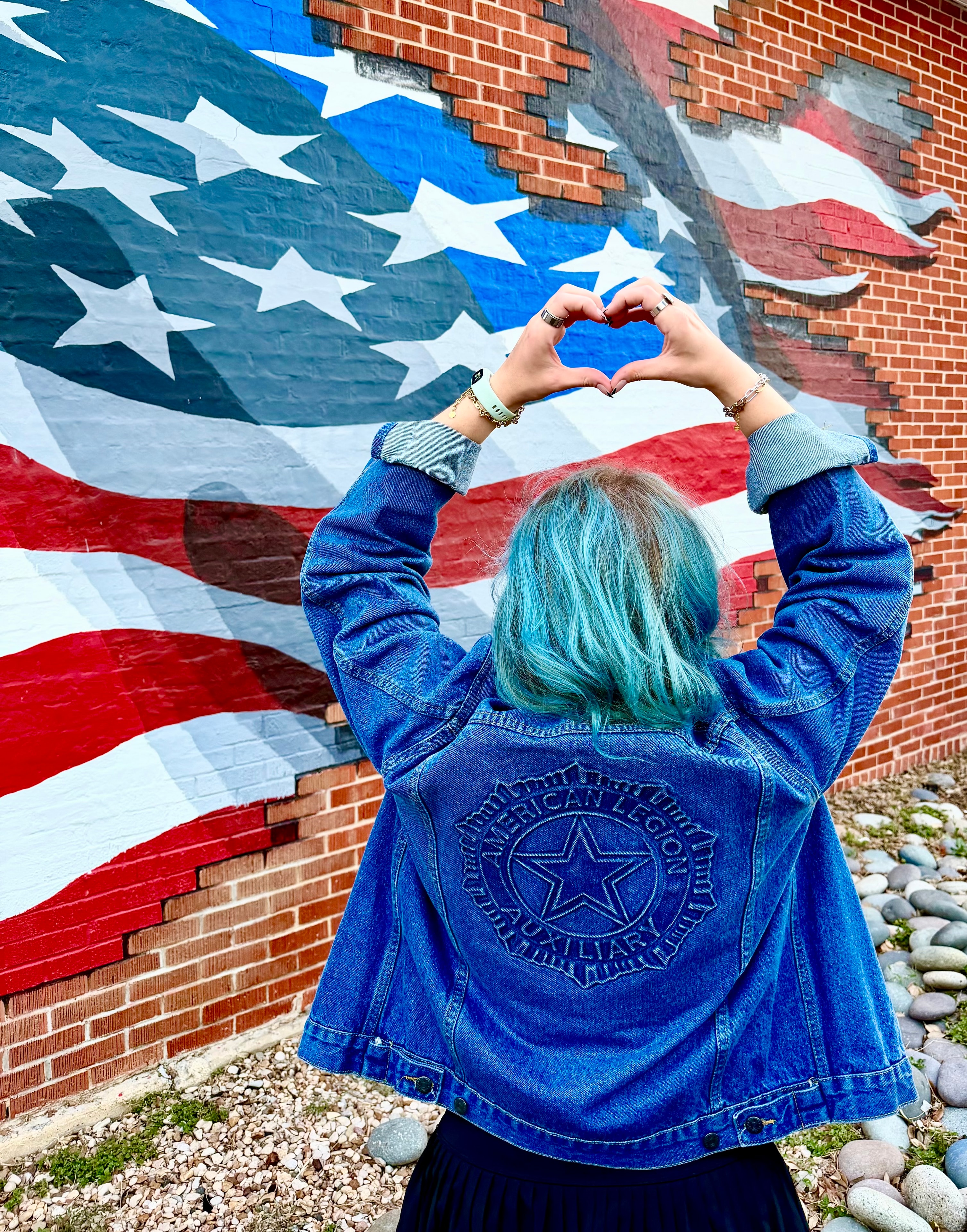 American Legion Auxiliary member making heart hands in front of American flag mural, ALA emblem jacket